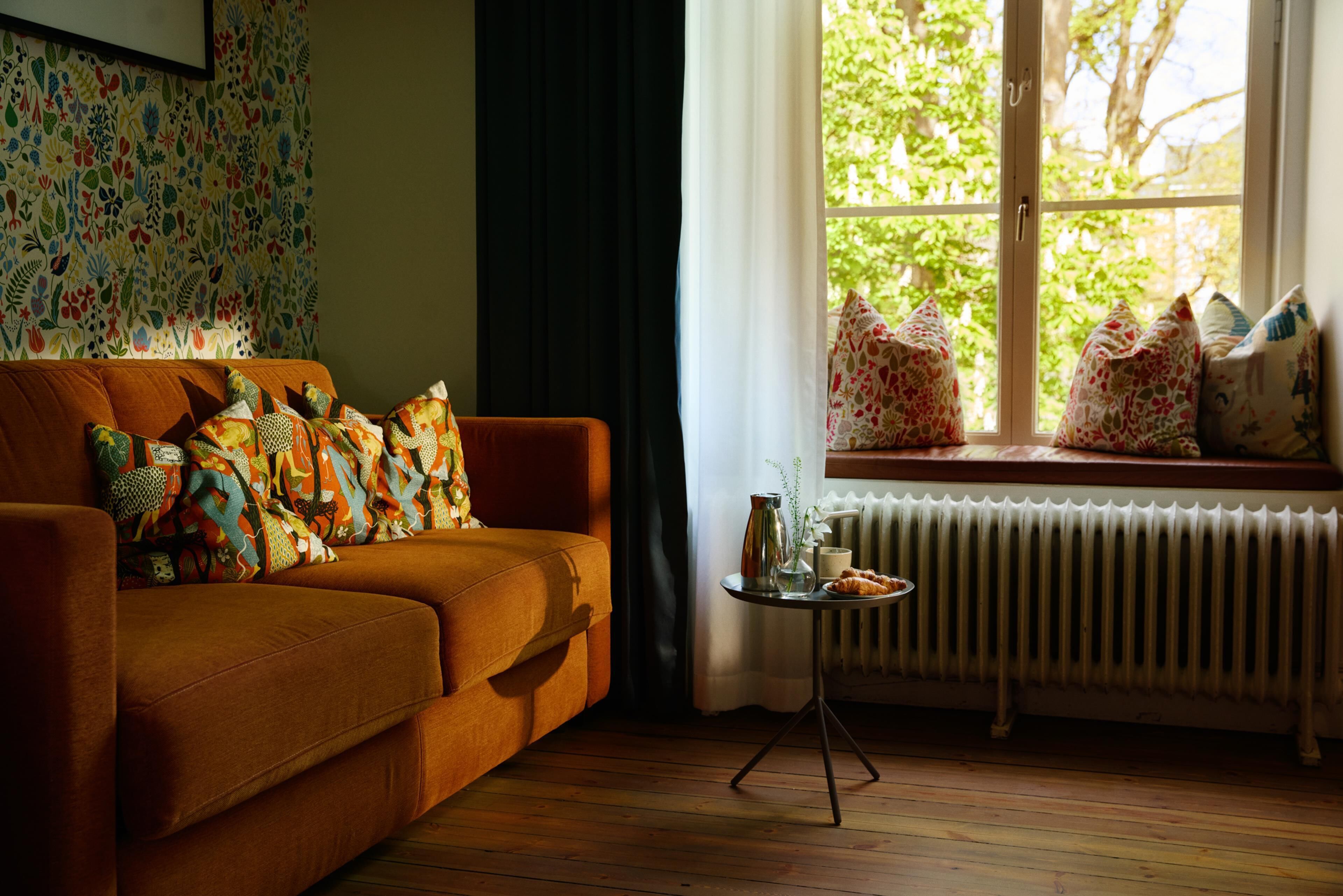 Un salon chaleureux avec un canapé, des coussins à motifs, une petite table sur laquelle se trouvent un vase et une tasse, et une fenêtre donnant sur un paysage verdoyant.