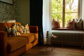 Una sala de estar acogedora con un sofá, cojines estampados, una mesita con un jarrón y una taza, y una ventana que da a vistas de vegetación.