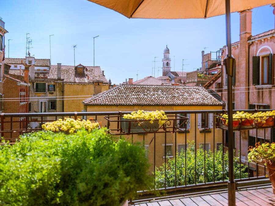 Uma varanda com grades de metal, plantas em vasos e vista para edifícios com telhado de azulejos e uma torre.