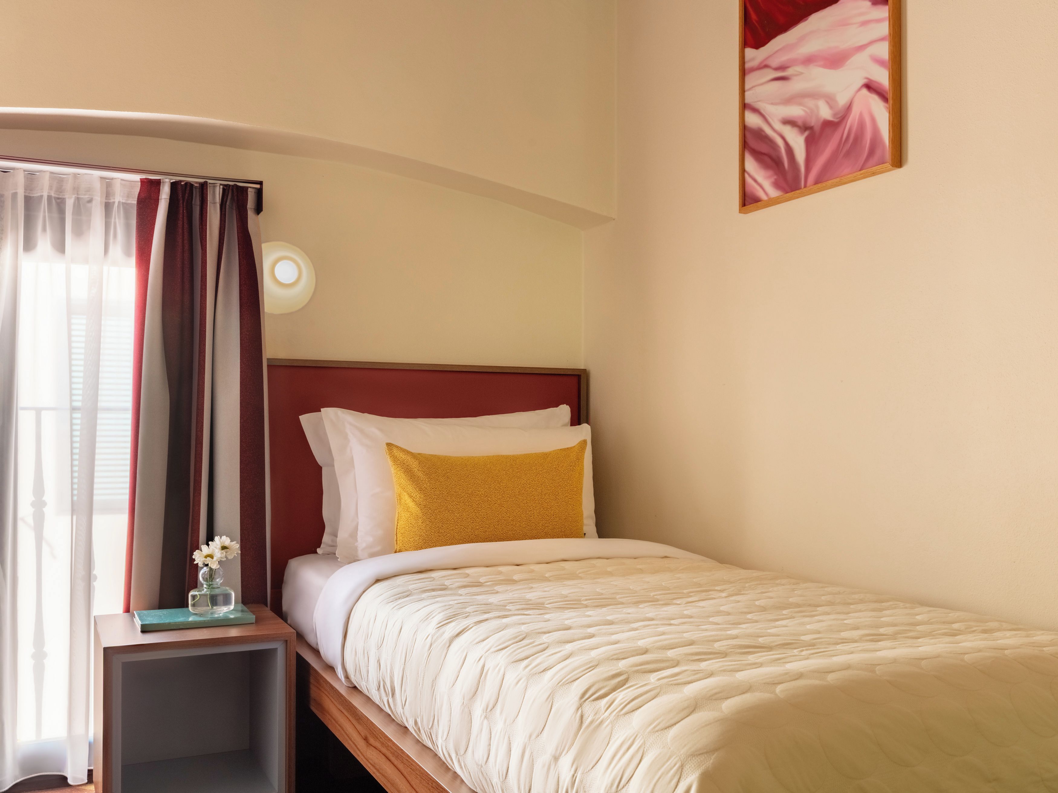 A single bed with a headboard, a bedside table with a lamp, and a window with curtains.