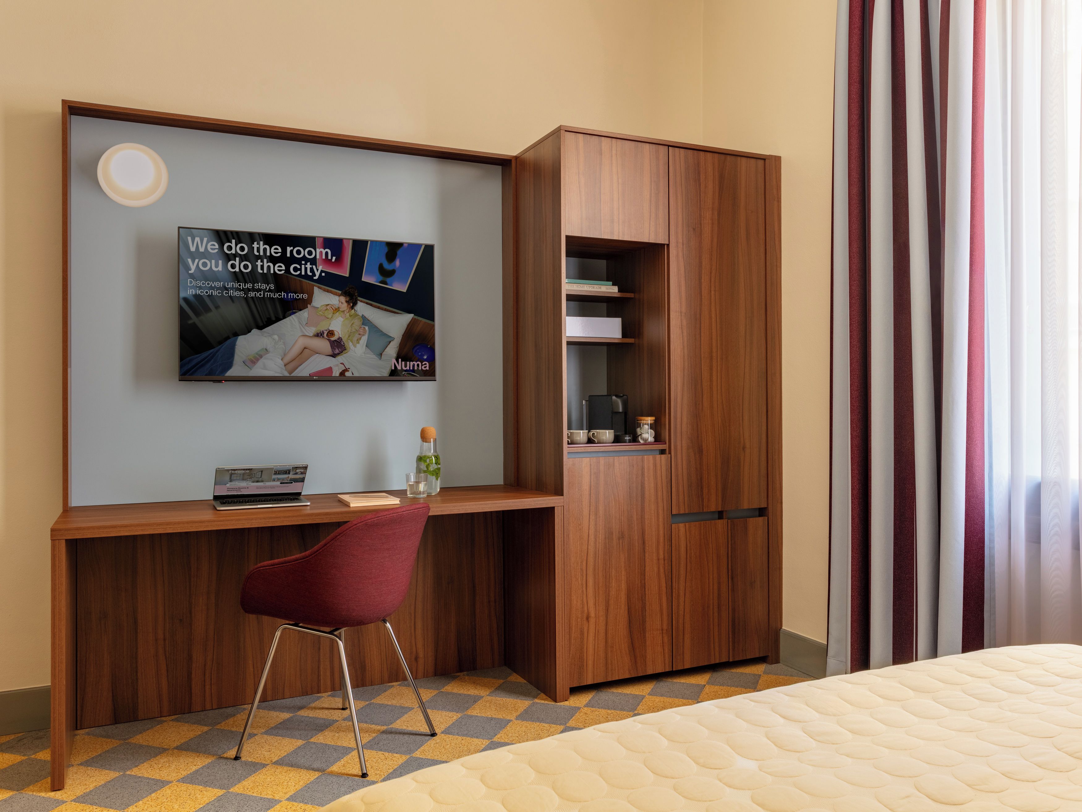 A hotel room with a wooden desk, a red chair, a bed with a quilted mattress, a television mounted on the wall, and a wardrobe with a mirror.