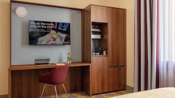A hotel room with a wooden desk, a red chair, a bed with a quilted mattress, a television mounted on the wall, and a wardrobe with a mirror.
