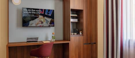 A hotel room with a wooden desk, a red chair, a bed with a quilted mattress, a television mounted on the wall, and a wardrobe with a mirror.