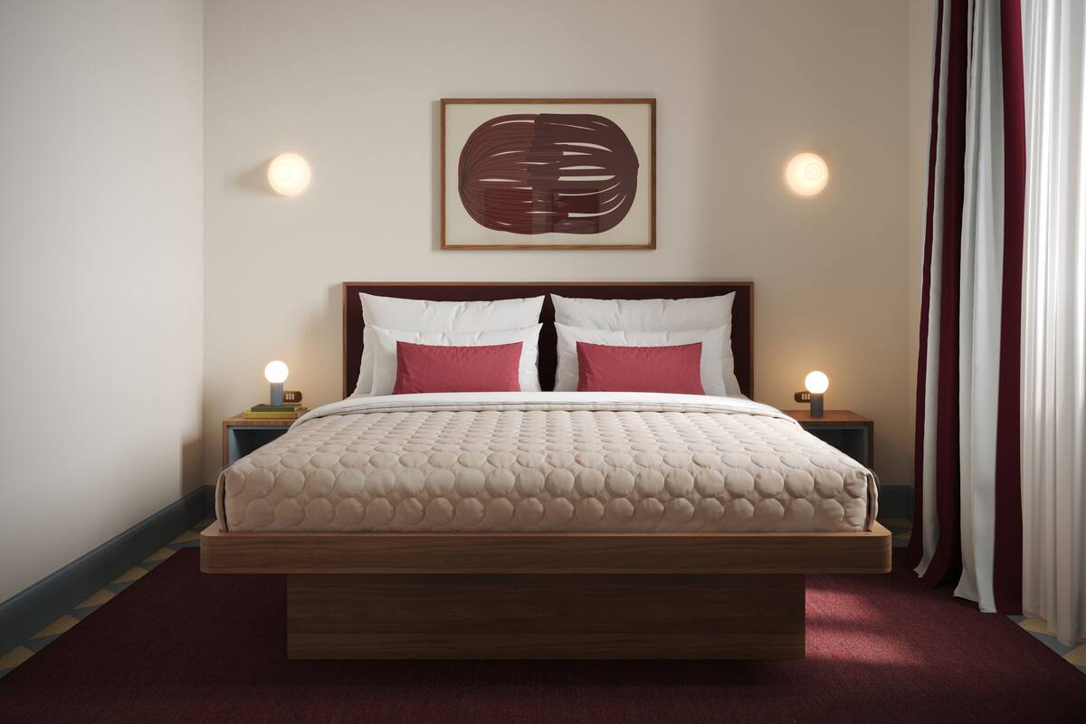 A bedroom with a bed, two bedside tables, a wall-mounted light, a framed artwork, and a window with curtains.