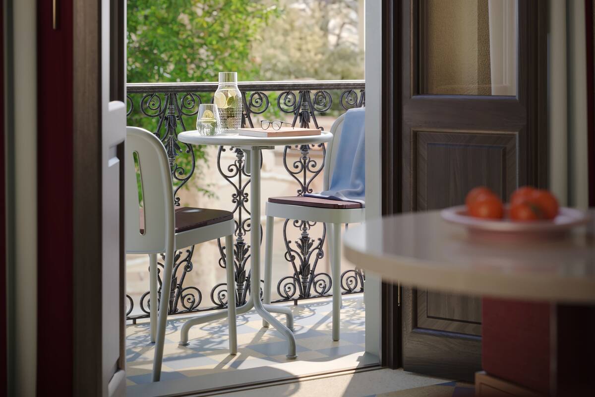An open door leading to a balcony with a table and chairs, a small plate with tomatoes, and a view of greenery outside.