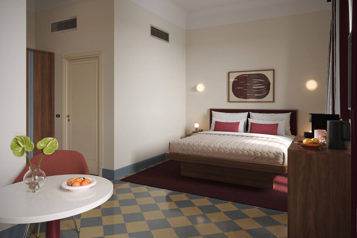 A hotel room with a bed, bedside table, a small table with a vase and fruit, and a checkered floor.