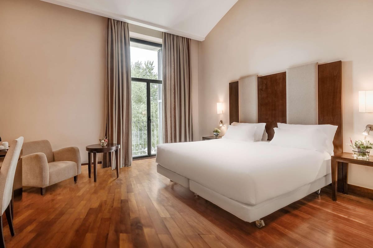 Habitación de hotel con una cama grande, un escritorio con silla, una mesita de noche con lámpara, y vistas al exterior a través de una ventana.