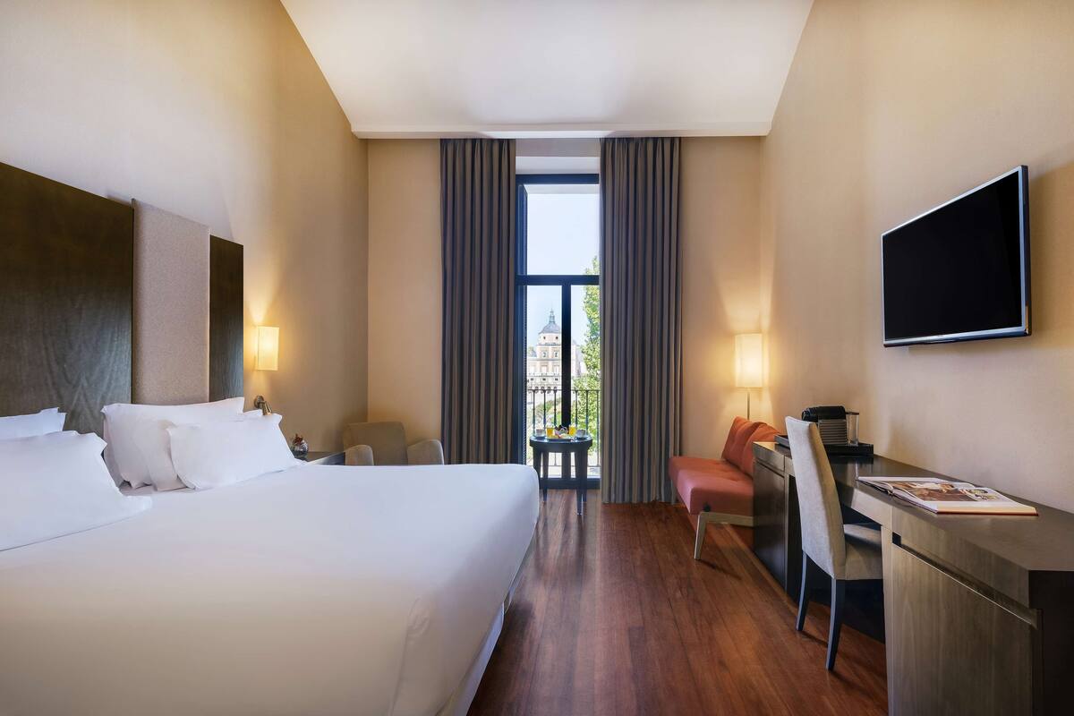 A hotel room with a large bed, a desk, a chair, a TV, and a view of a building through the window.