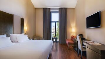 A hotel room with a large bed, a desk, a chair, a TV, and a view of a building through the window.