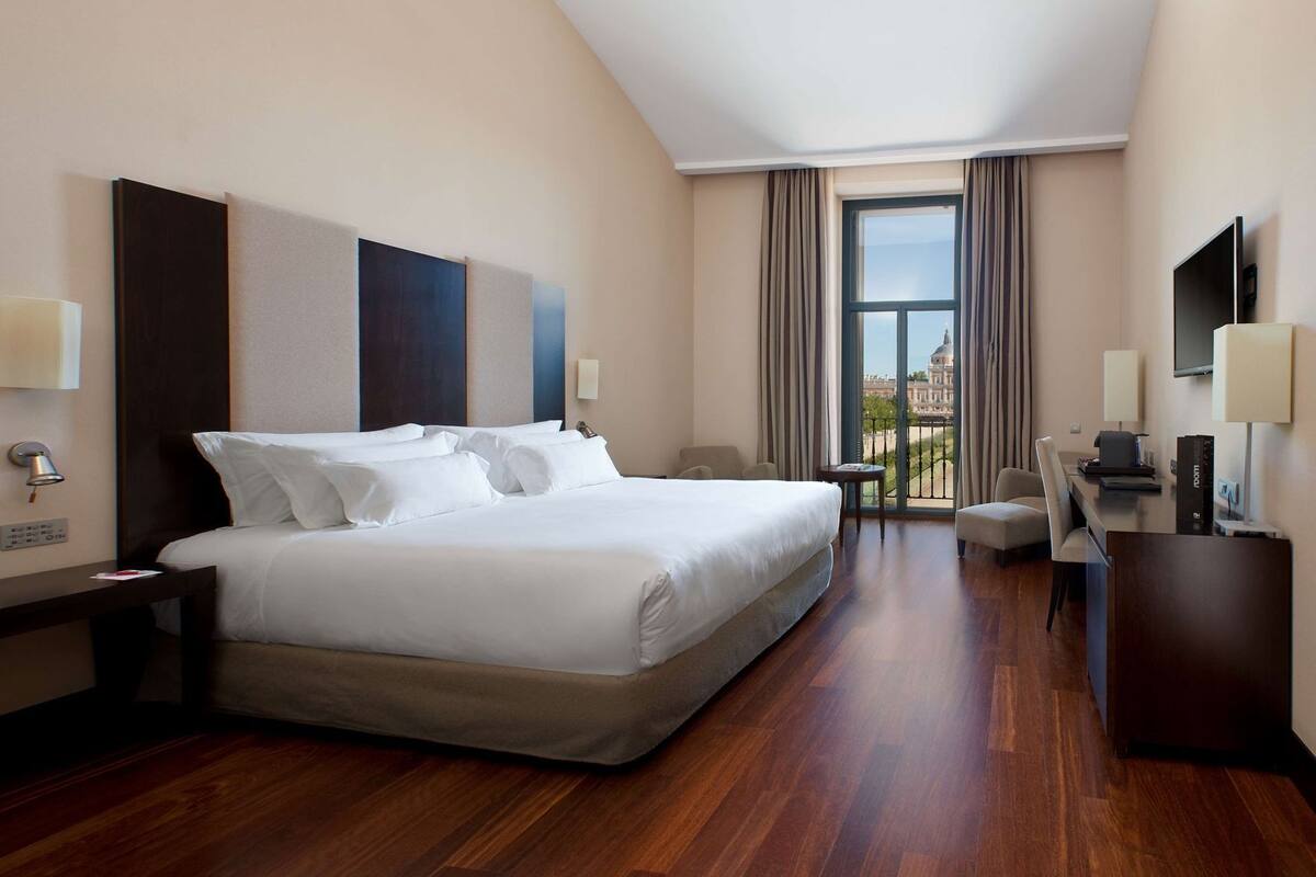 A hotel room with a large bed, a desk, a chair, a TV, and a view of a building through the window.