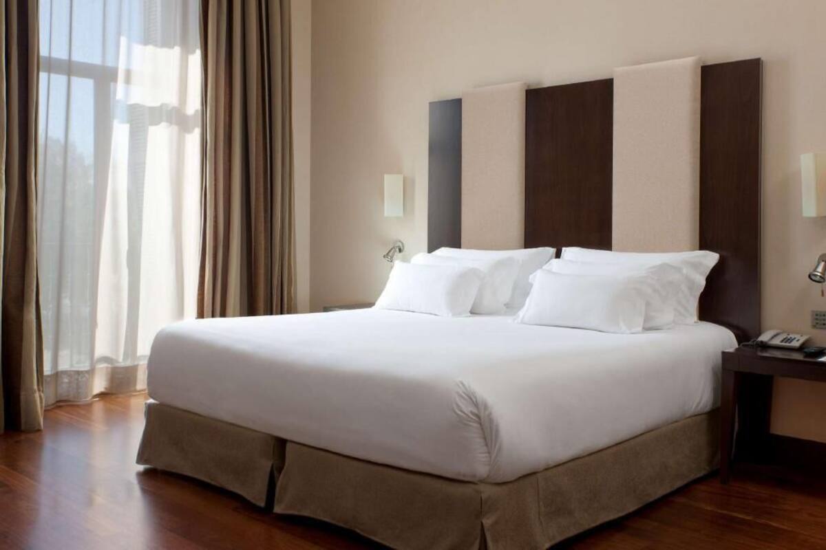 A hotel room with a large bed, a bedside table with a lamp, a window with curtains, and a wall-mounted telephone.