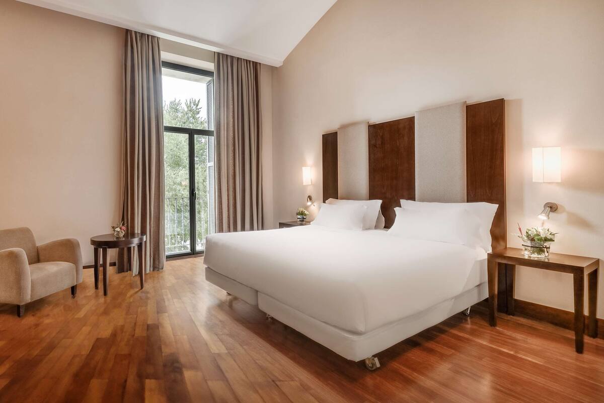 A hotel room with a large bed, a desk with a chair, a small table with a lamp, and a view through the window.