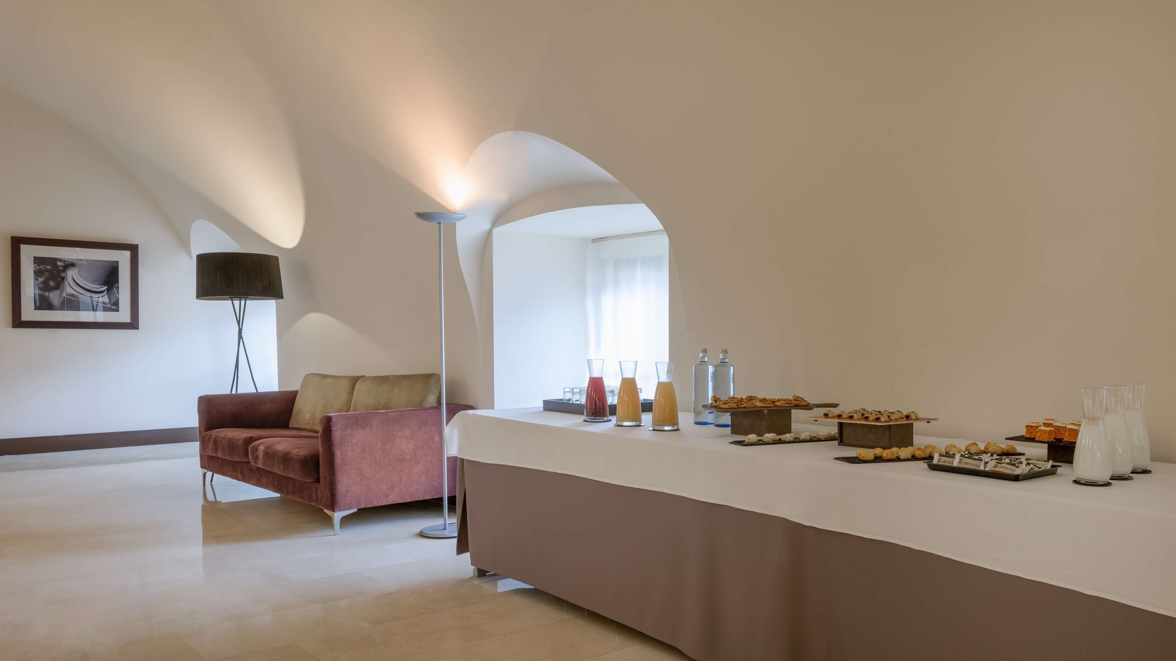 A modern hotel room with a long counter displaying food and drinks, a sofa, a floor lamp, and a framed picture on the wall.