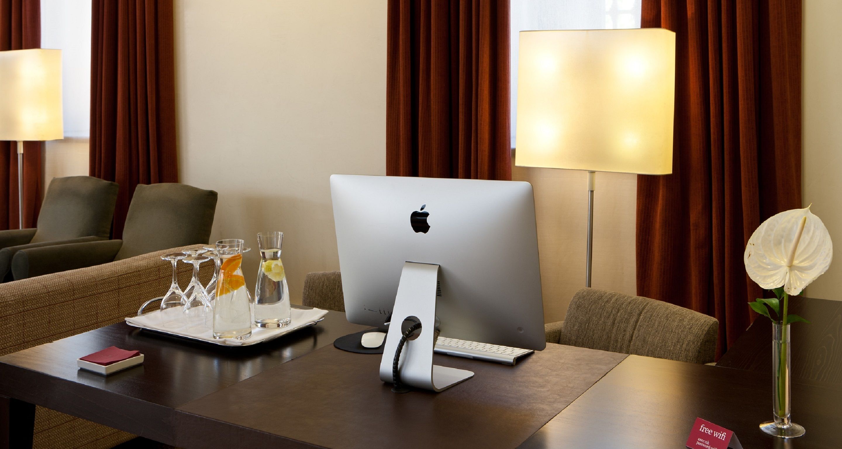 Habitación de hotel con un escritorio que incluye un iMac de Apple, una lámpara, un florero con una flor blanca y una bandeja con vasos y una botella.