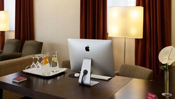 Habitación de hotel con un escritorio que incluye un iMac de Apple, una lámpara, un florero con una flor blanca y una bandeja con vasos y una botella.