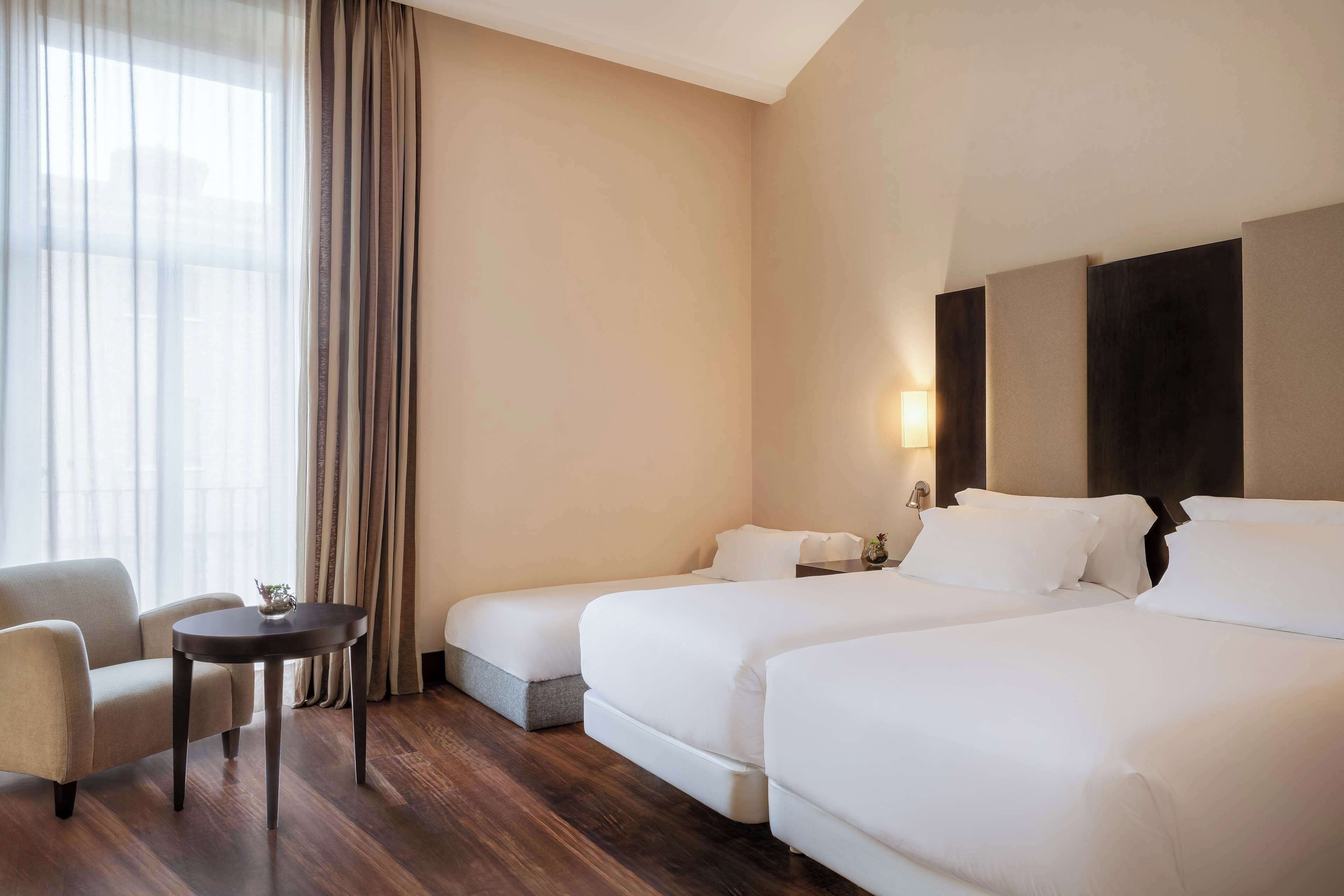 Habitación de hotel con dos camas, un sofá, una mesita y una ventana con cortinas.