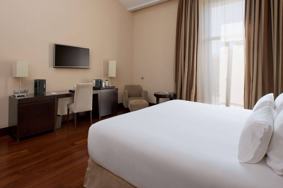 A hotel room with a large bed, a desk with a chair, a TV, and a window with curtains.