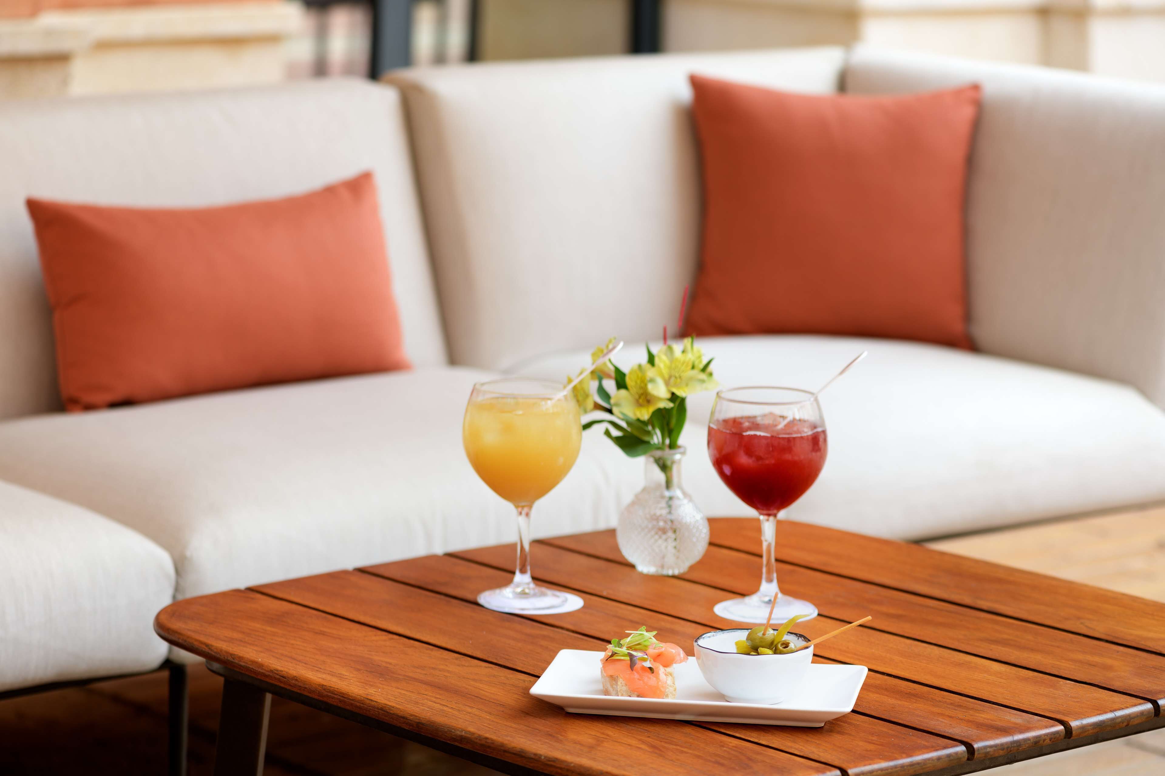 Une table en bois sur laquelle sont posés deux verres et une petite assiette de nourriture, le tout placé sur un canapé blanc agrémenté de coussins orange.