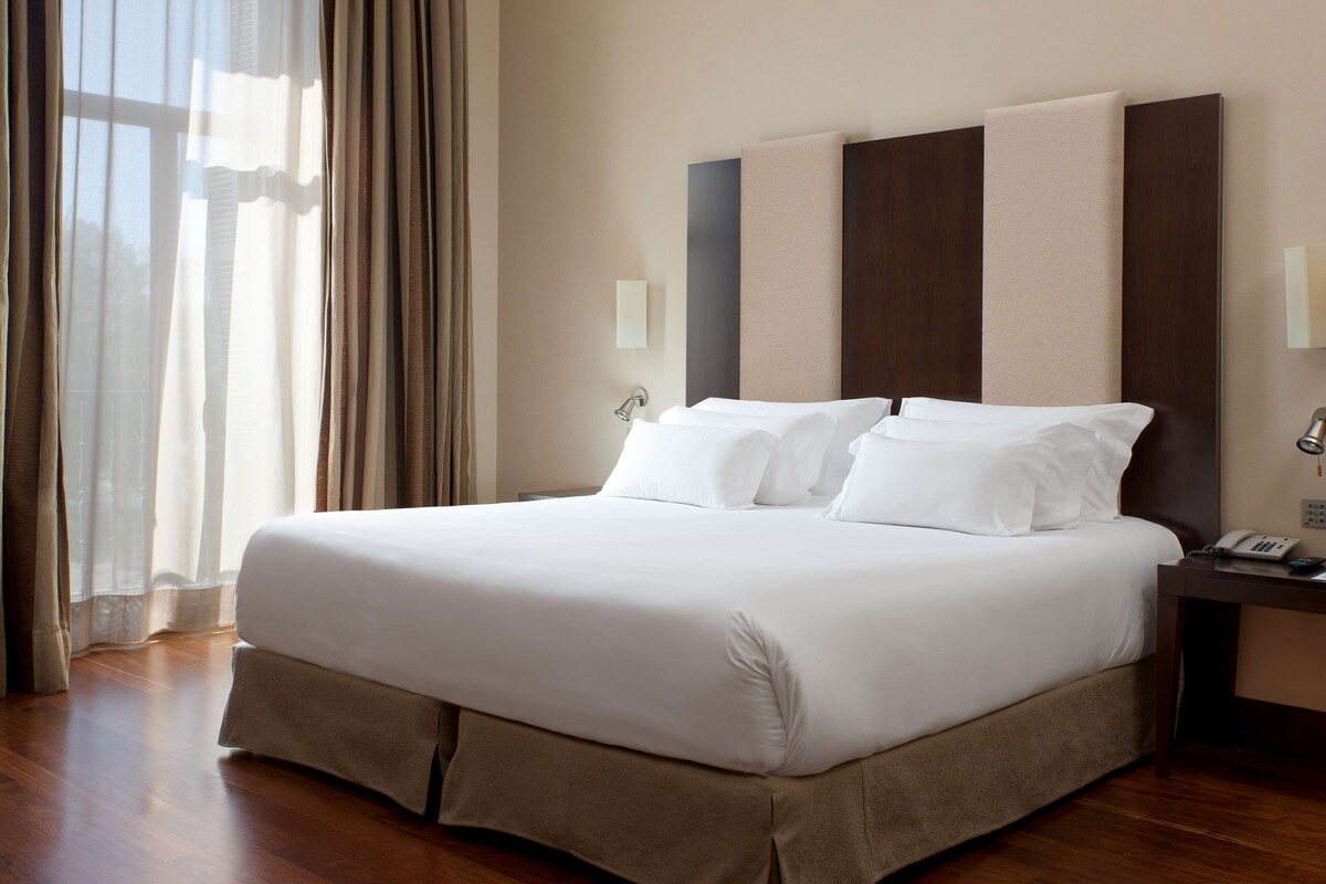 A hotel room with a large bed, a nightstand with a phone, and a window with curtains.