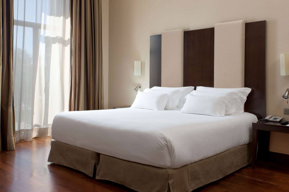 A hotel room with a large bed, a bedside table with a lamp, and a window with curtains.