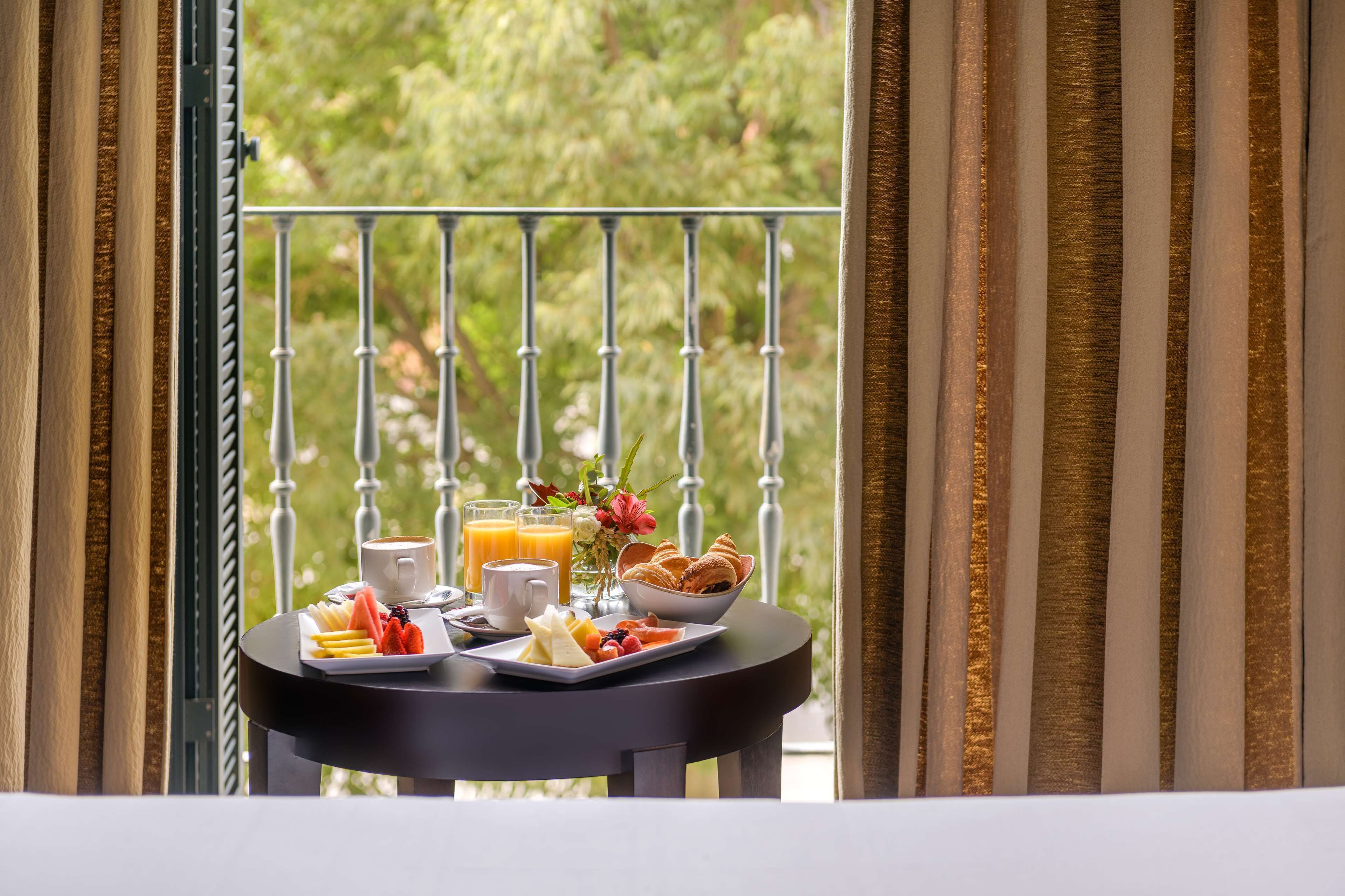 Une table de petit-déjeuner dressée avec une variété d’aliments et de boissons, placée devant un balcon offrant une vue sur la verdure.