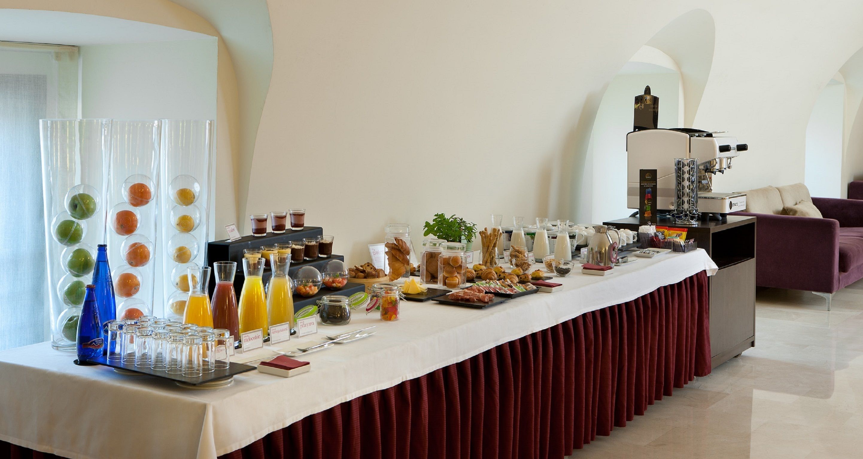 Una mesa de buffet con diversos alimentos y bebidas, una máquina de café y un área de estar con un sofá.
