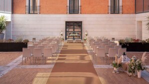 Un espacio para ceremonia al aire libre con filas de sillas y un pasillo alfombrado, flanqueado por arreglos florales y faroles decorativos.