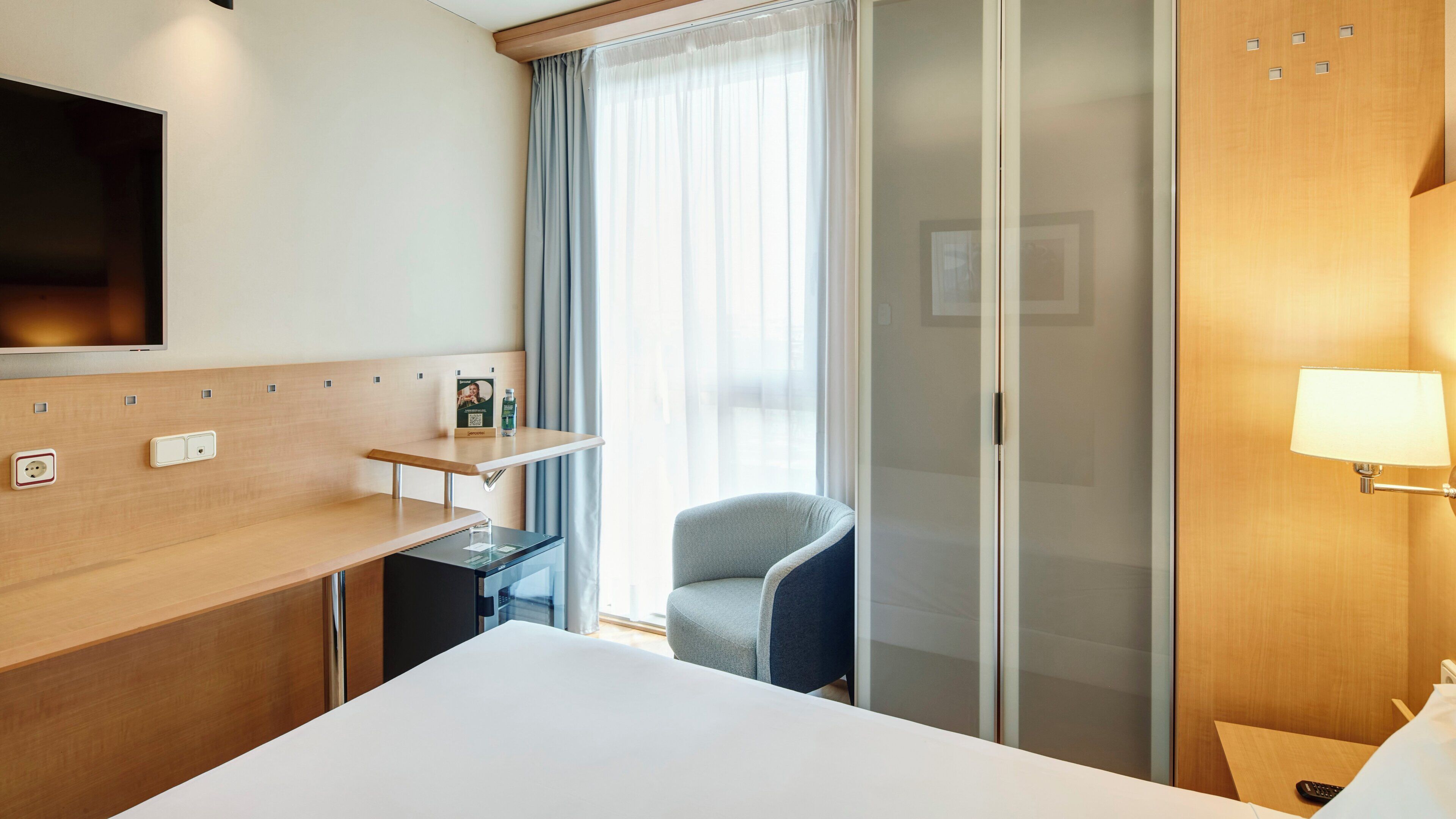 A hotel room with a bed, a desk, a chair, a TV, a lamp, and a window with curtains.