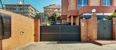 A gated entrance with a brick wall, a closed metal gate, and a building with a red facade in the background.