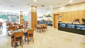 A hotel dining area with multiple tables and chairs, a buffet counter with food items, and a view of the outdoors through large windows. - Sercotel Porta Barcelona (Sant Just Desvern)