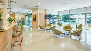 A hotel lobby with a reception desk, seating area, and large windows offering a view of the outdoors. - Sercotel Porta Barcelona (Sant Just Desvern)
