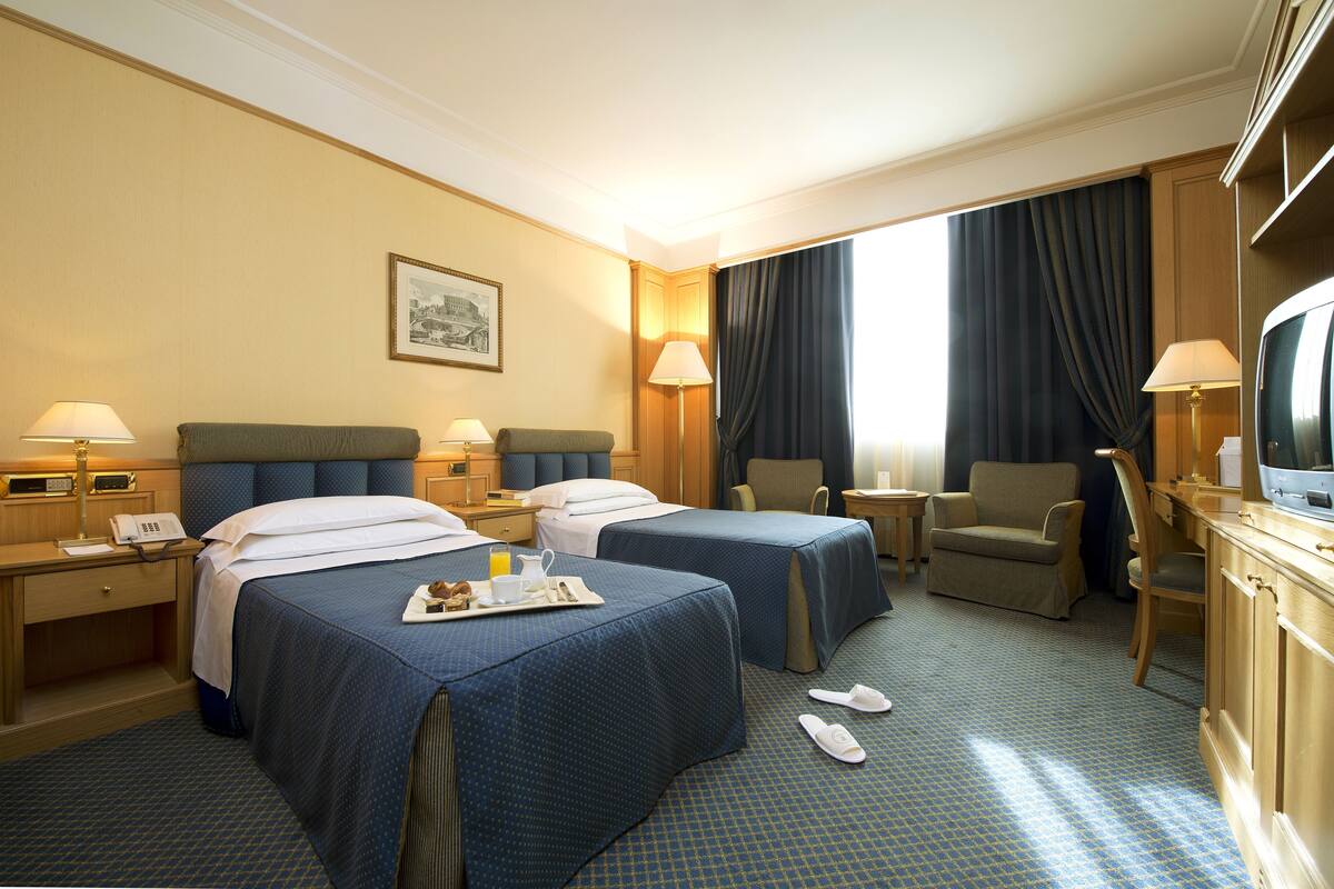 A hotel room with two beds, a desk, a TV, and a chair.