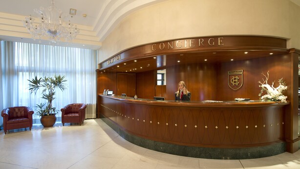 Een hotel receptie met een gebogen balie, een kroonluchter, twee leren fauteuils en een potplant.