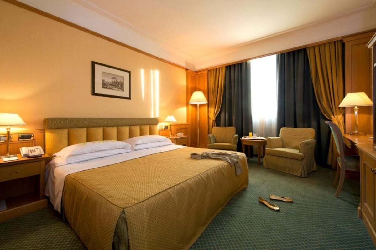 Habitación de hotel con una cama grande, dos mesitas de noche con lámparas, una silla, un escritorio con televisor y una ventana con cortinas.