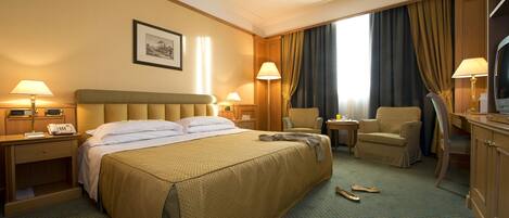 Una habitación de hotel con una cama grande, dos mesitas de noche con lámparas, una silla, un escritorio con silla, un televisor y una ventana con cortinas.