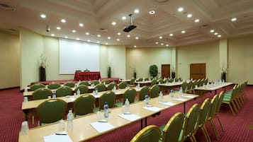 Una sala de conferències amb files de cadires verdes, taules amb ampolles d'aigua i una pantalla de projector.