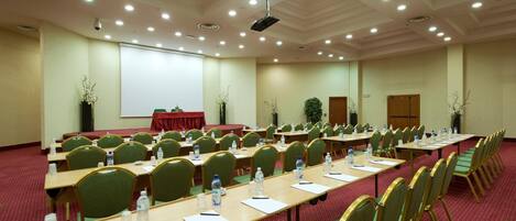 A conference room with rows of green chairs, tables with water bottles, and a projector screen.