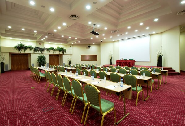 Un salón de conferencias con filas de sillas verdes y mesas, una pantalla de proyector y un mantel rojo.