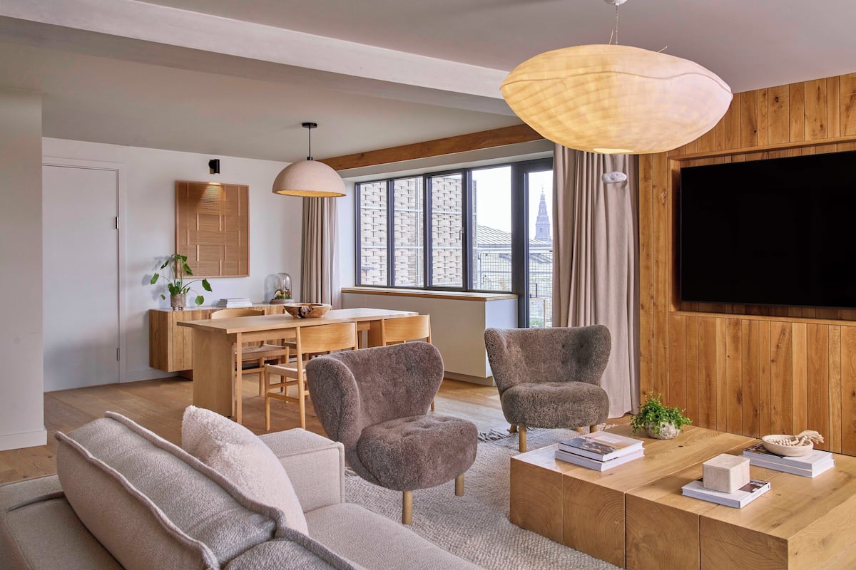 A modern living room with a sofa, armchairs, a coffee table with books, a TV, and a dining area with a wooden table and chairs.