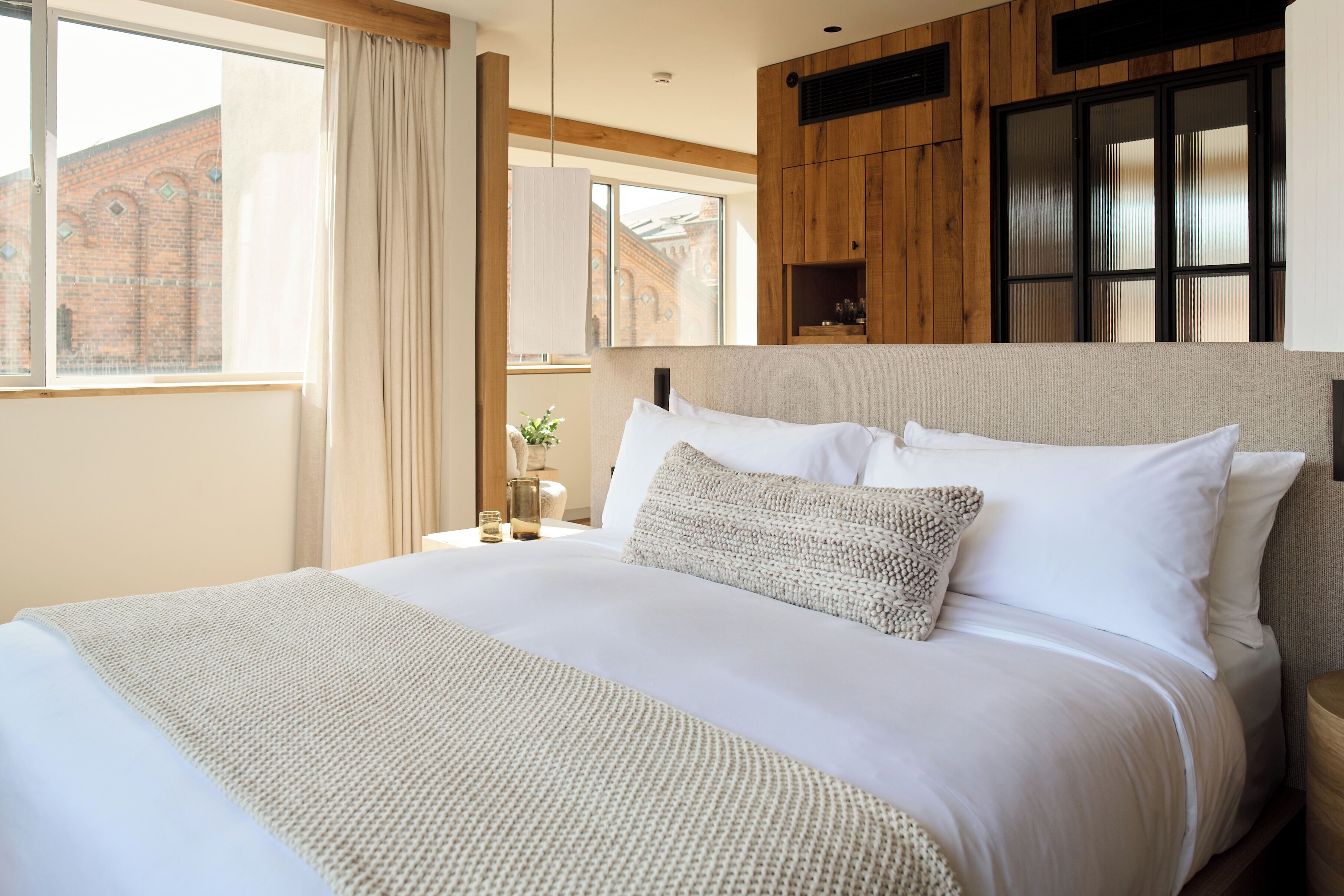 A neatly made bed with white linens and a textured throw blanket, a wooden headboard, and a view of a brick building through a window.