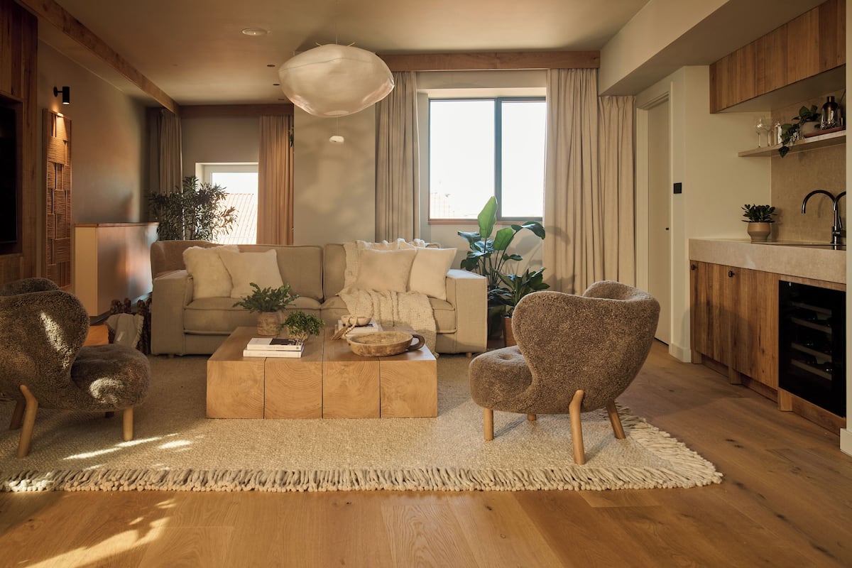 A modern living room with a sofa, coffee table, and armchairs, a wooden floor, and a large window with curtains.