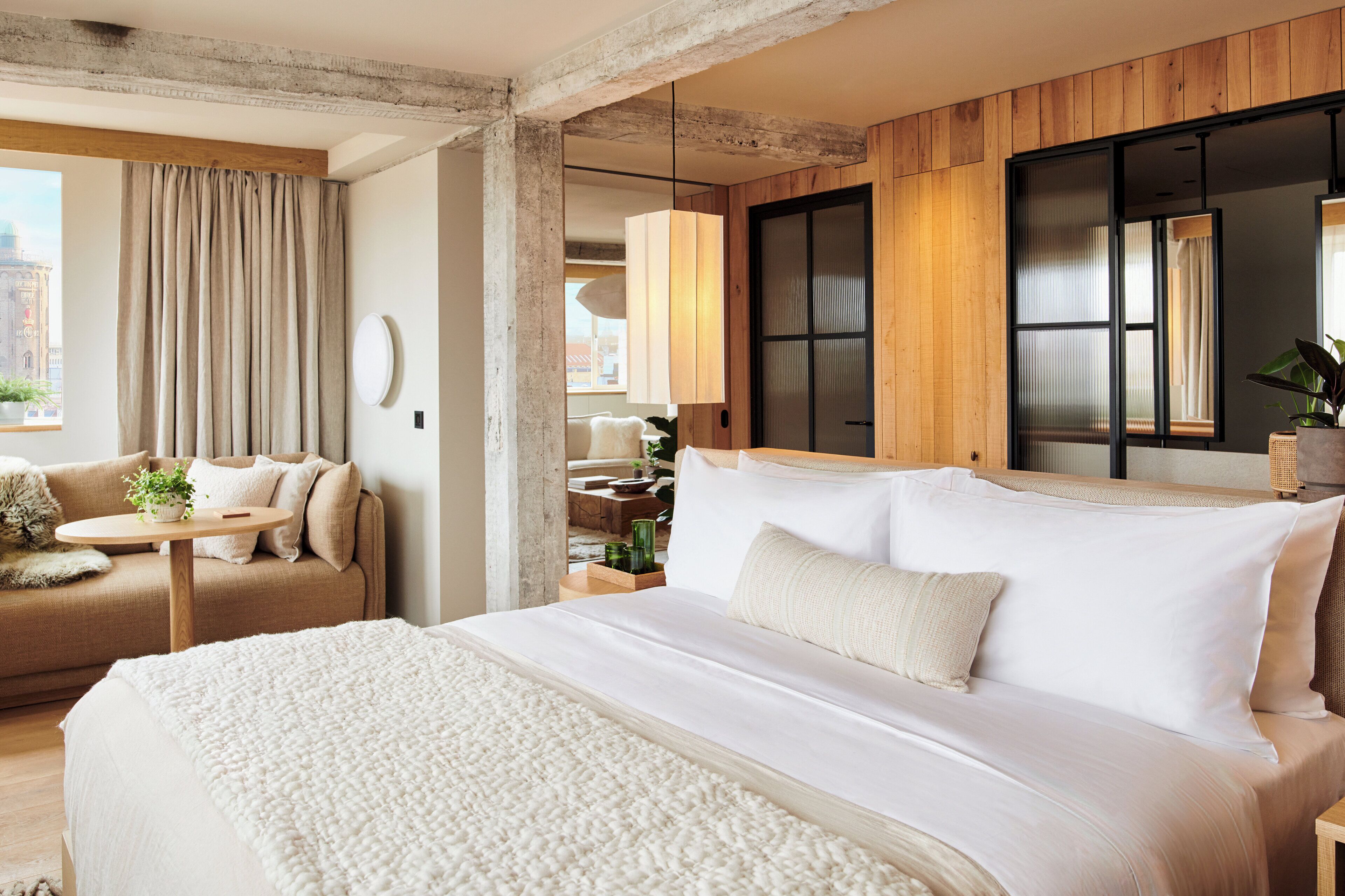 A modern hotel room with a large bed, a sofa, a small table with a plant, and a view of the city.