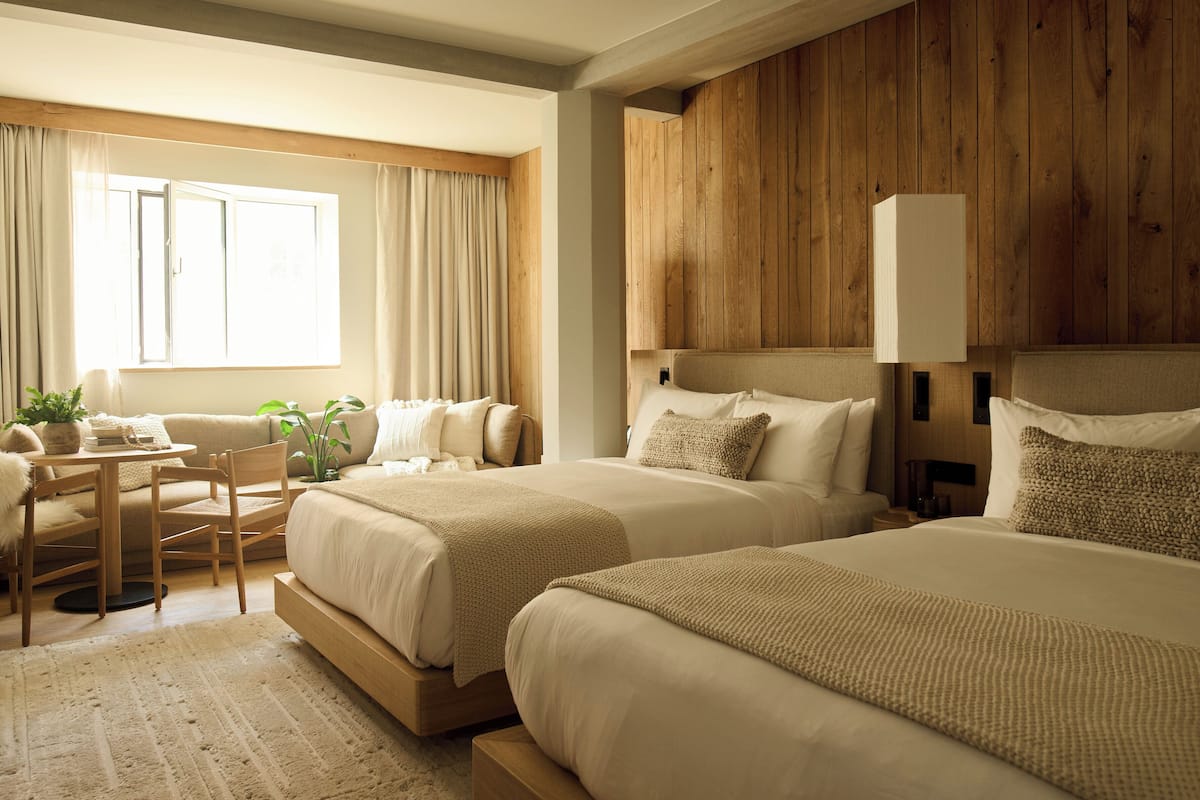 A hotel room with two beds, a wooden headboard, a small table with chairs, and a window with curtains.
