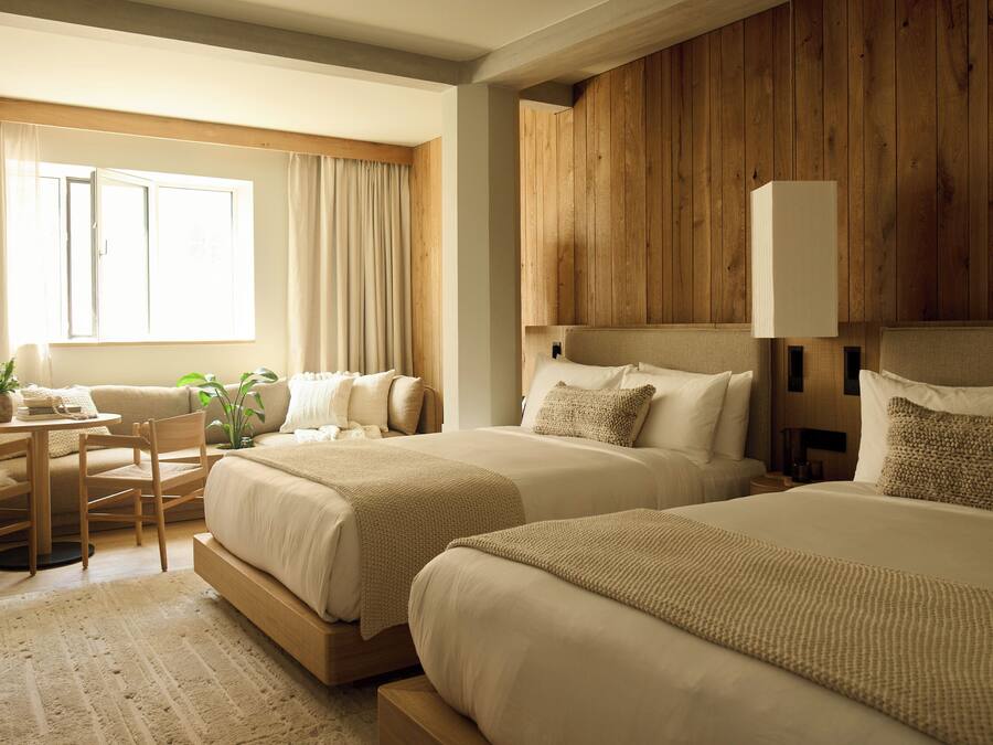 Habitación de hotel con dos camas, cabecero de madera, una mesita con sillas y una ventana con cortinas.