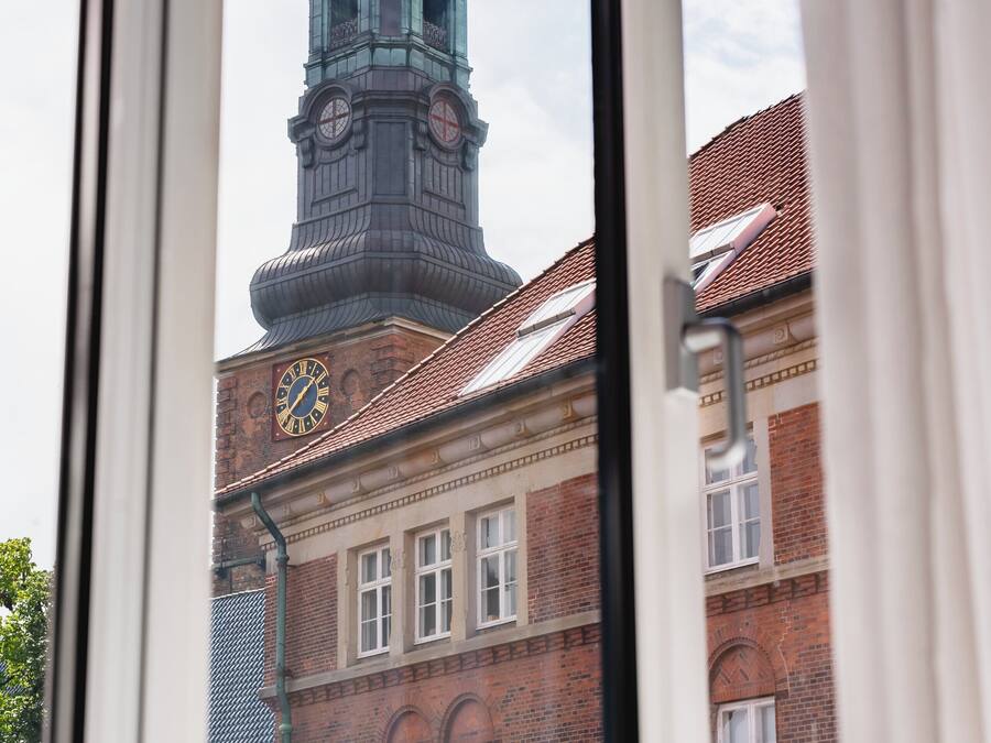 A window view of a building with a clock tower, a brick facade, and a window with a curtain.