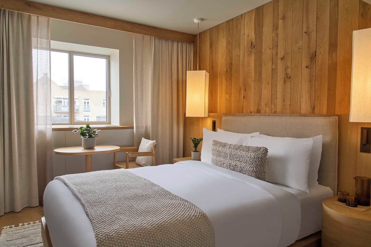A hotel room with a large bed, a small round table, a chair, a window with curtains, and wooden paneling on the wall.