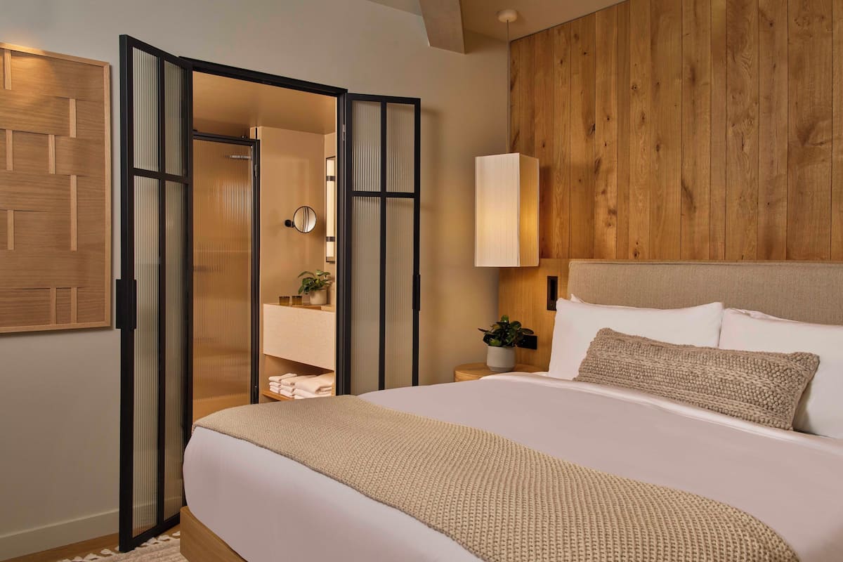 A bedroom with a large bed, a wooden headboard, a nightstand with a lamp, a mirror, and a sliding door leading to a bathroom.