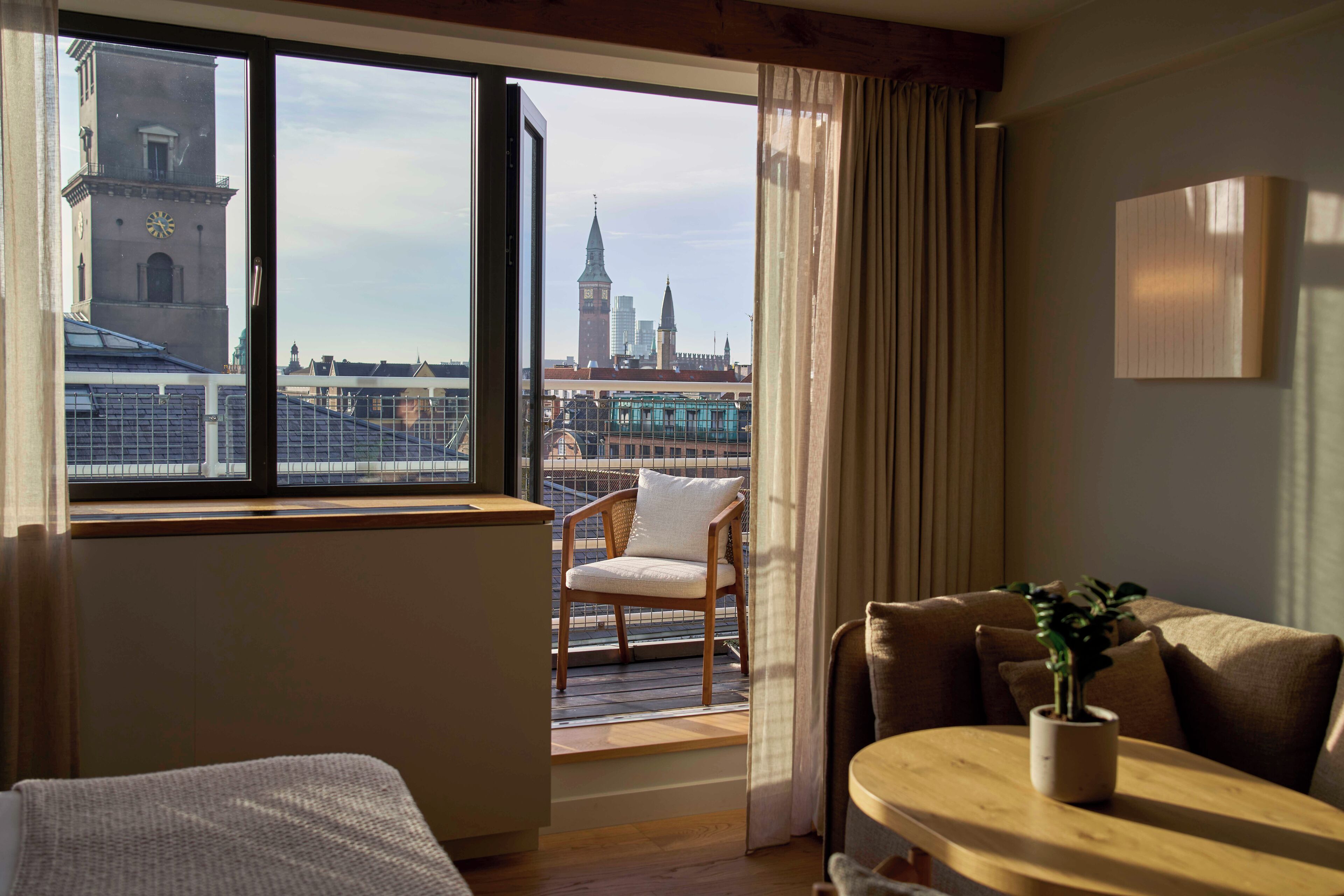 A hotel room with a balcony, a view of a city skyline, a wooden chair, a sofa, and a small table with a plant.
