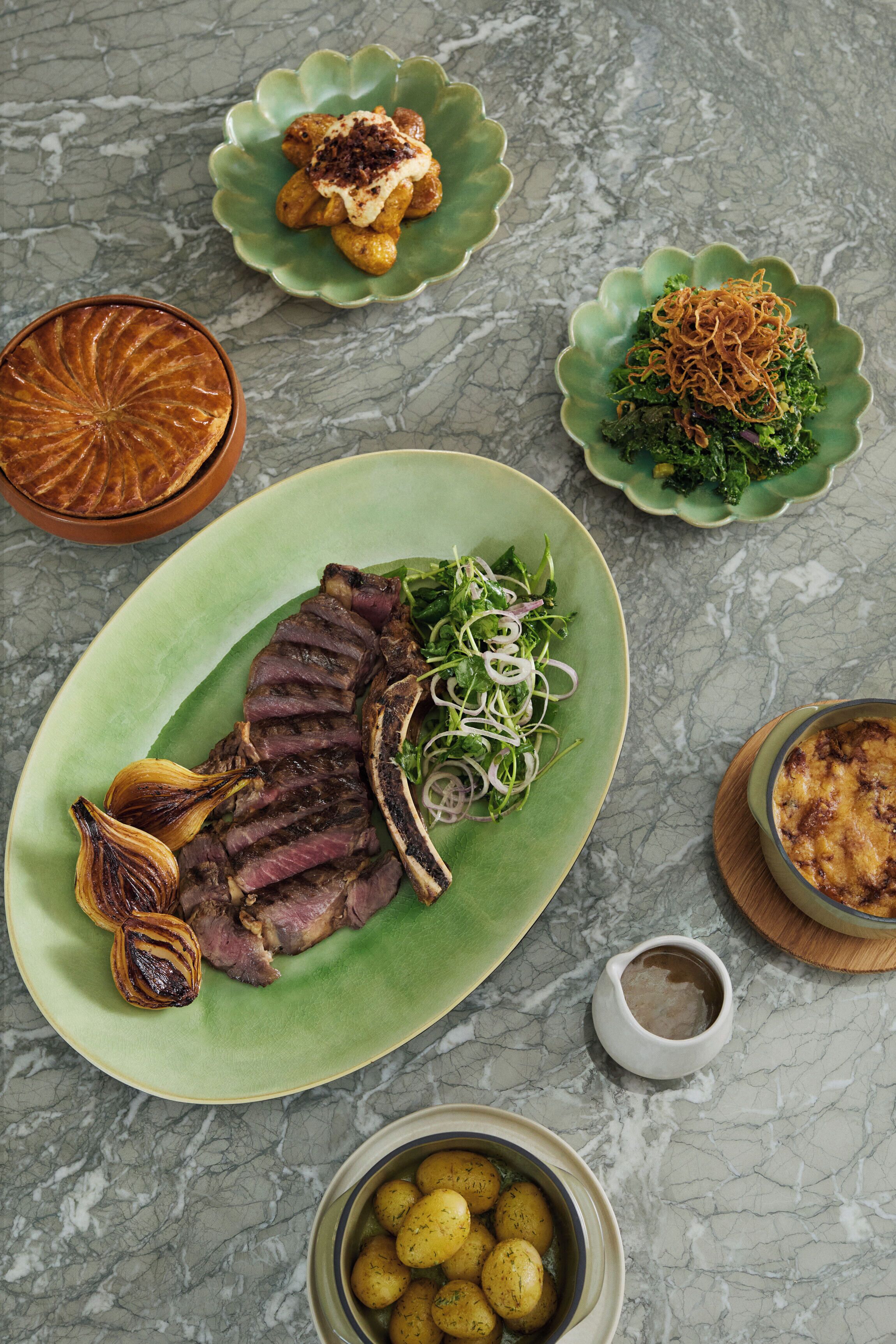 Ein Tisch mit Marmorplatte und verschiedenen Gerichten, darunter ein Steakteller, Bratkartoffeln und eine Schüssel mit Salat.