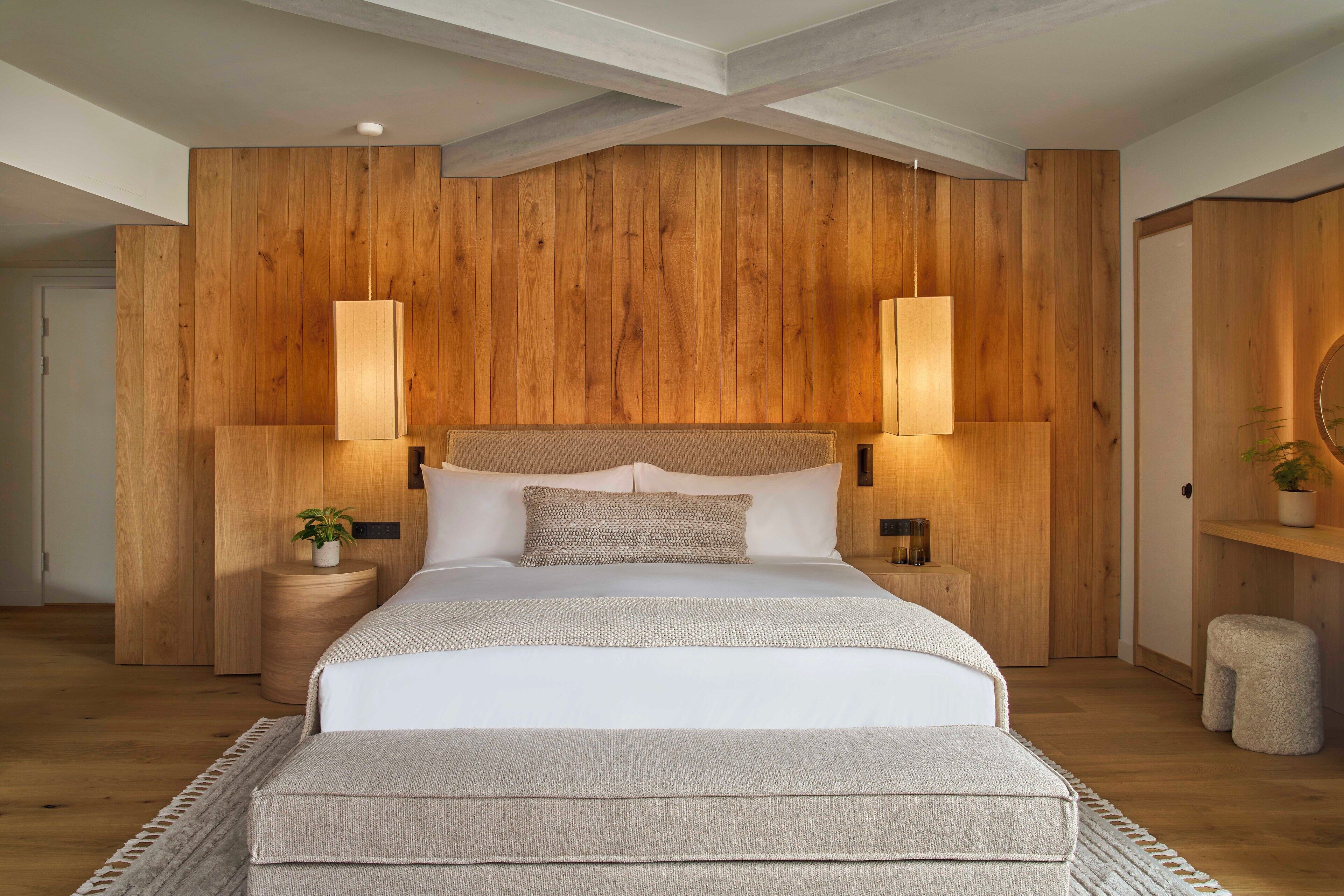 A bedroom with a wooden headboard, two bedside lamps, a bed with white bedding, a small bedside table with a plant, and a round mirror on a wooden shelf.
